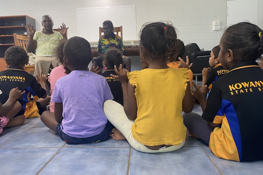 School children sit on a tiled floor with their hands in the air while singing songs with Aboriginal elders.