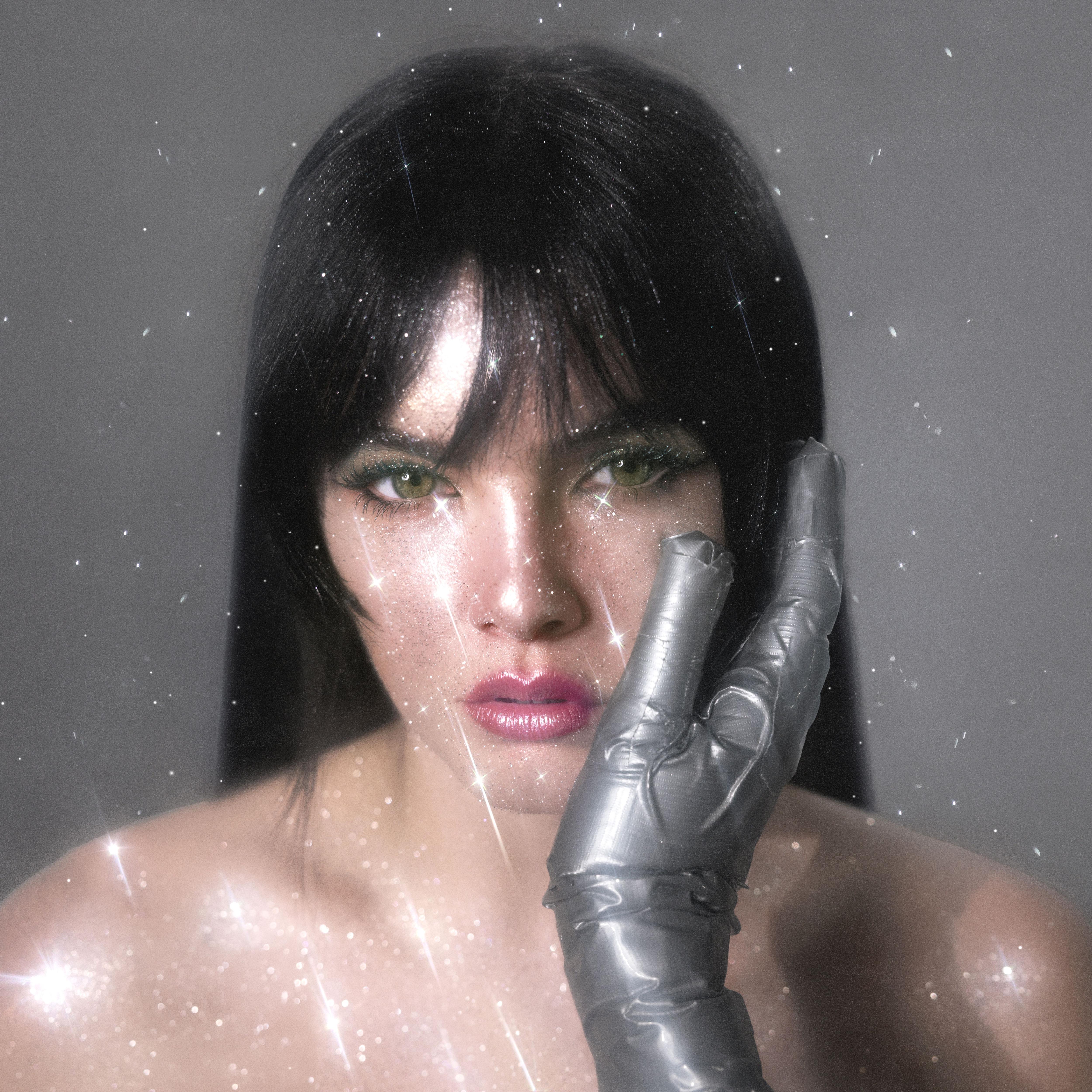 Dark hair female with fridge looking into camera. A silver gloved hand is on her face. Sparkles visible over whole image