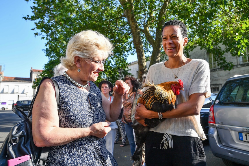Corinne Fesseau stands next to a woman holding her rooster Maurice.