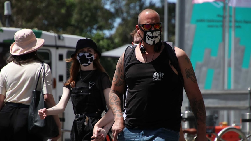 A man in a singlet and a Guy Fawkes-inspired face mask walks in the sun, a woman in a cow-print mask behind him.