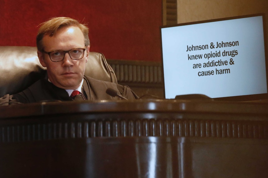 A judge listens during opening arguments for the trial with a screen next to him.