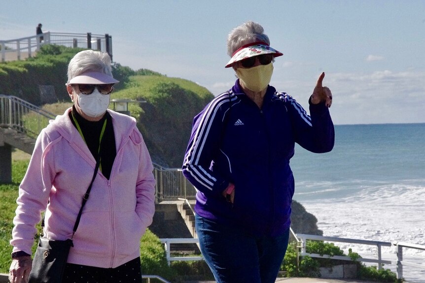 Two women wearing masks and tracksuits walk along a cliff top near a beach.