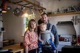 A young couple and their baby who is strapped to the father's chest look at the camera. The are standing in the kitchen.