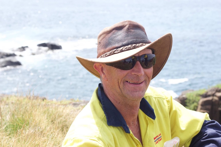 An aboriginal man wearing reflective sunglasses and a wide hat beams into the camera with an ocean landscape behind.