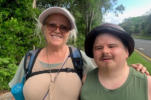 A middle-aged caucasian woman stands with her 39 year old son by a road, smiling at the camera, with a road in the background.