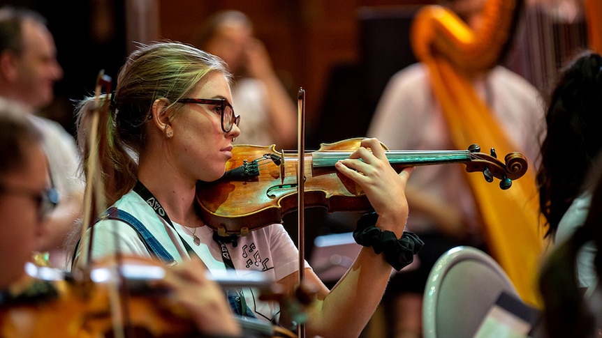 A student plays the violin with and out of focus orchestra in the background.