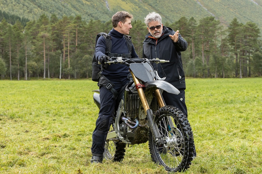 A man sits on a motorbike next to a man who is standing talking to him and pointing straight ahead.