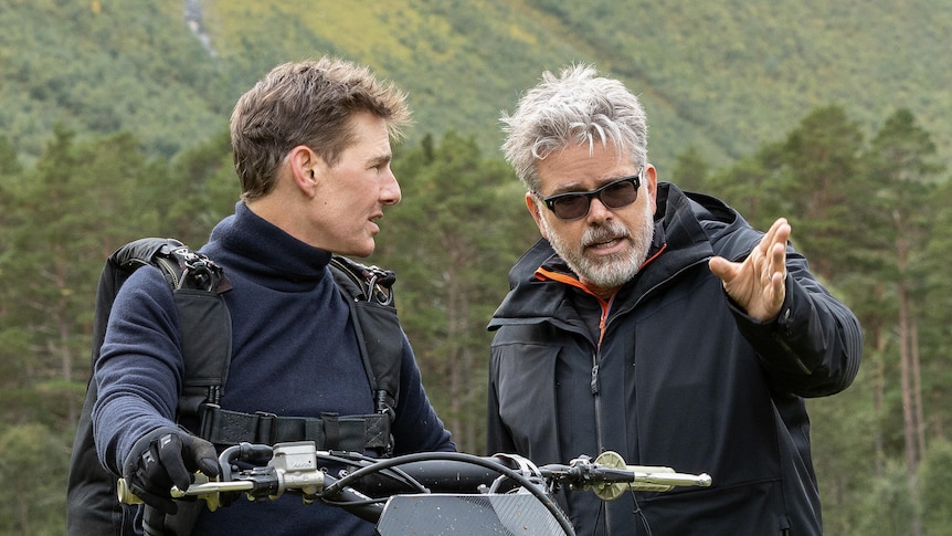 A man sits on a motorbike next to a man who is standing talking to him and pointing straight ahead.