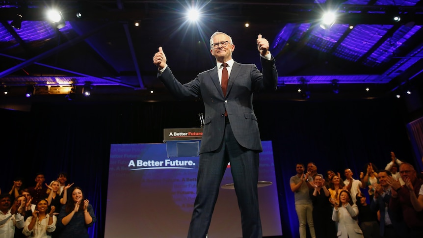 Albanese gives a thumbs up from a stage as an audience applauds him, with campaign slogans visible behind him.