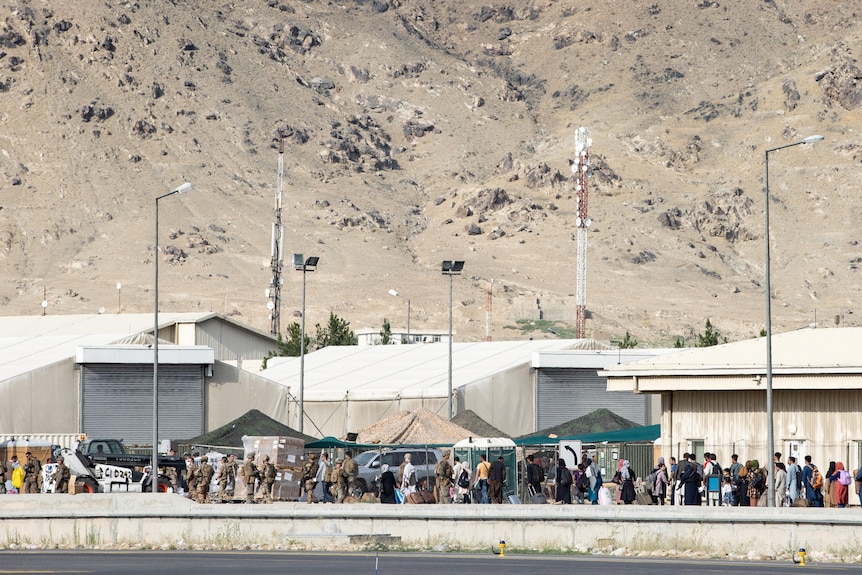 A wide shot shows hundreds of people waiting beside pop up tents outdoors at an airport.