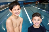 Two young boys standing in front of an indoor laned swimming pool with big smiles on their faces.