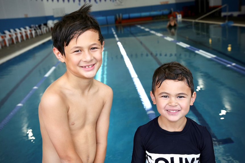 Two young boys standing in front of an indoor laned swimming pool with big smiles on their faces.