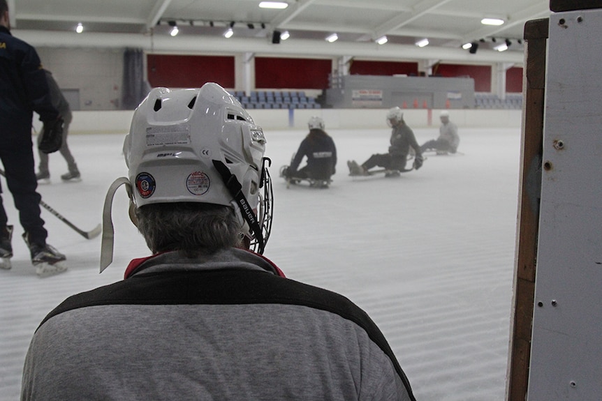 The back of a ice sledge hockey players helmet with people on sleds on an ice skating rink in the background.