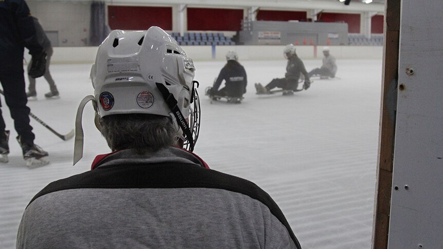 The back of a ice sledge hockey players helmet with people on sleds on an ice skating rink in the background.