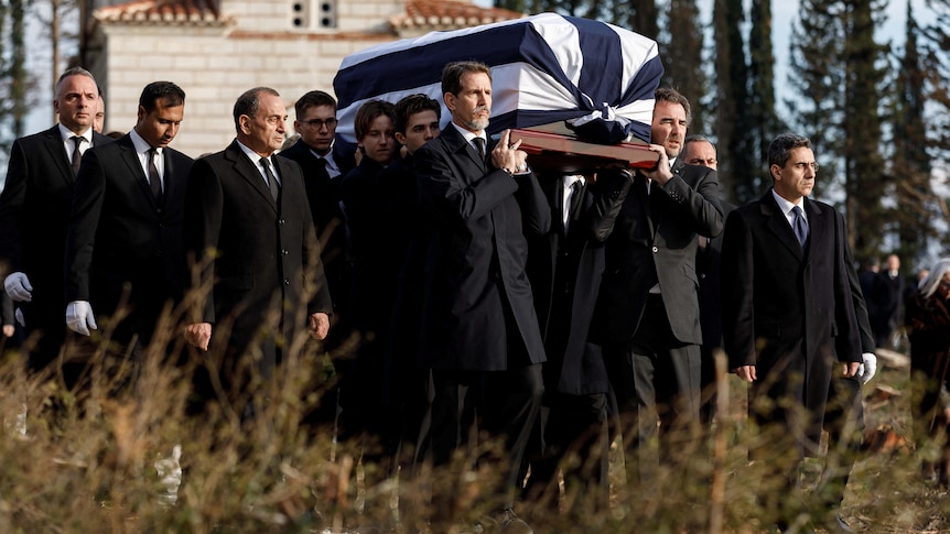 Men in black are pictured carrying a coffin that is draped in a Greek flag. In the background is greenery.