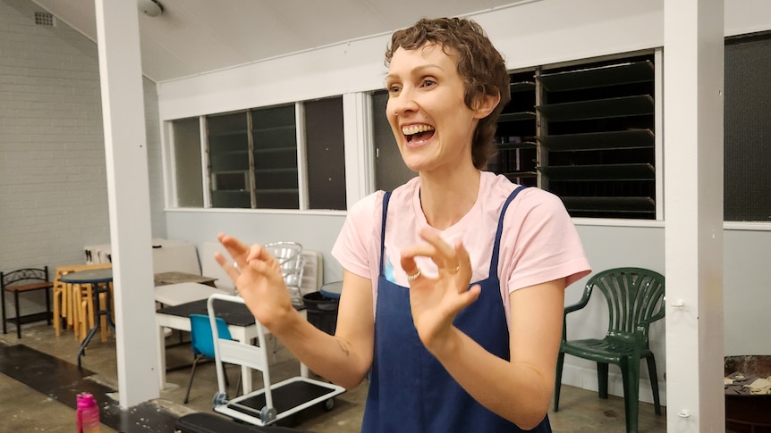 A woman wearing blue overalls over a pink shirt smiles while conducting, in a hall at night time