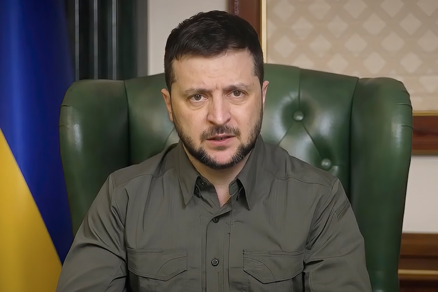 Volodymyr Zelenskyy sits in a chair in front of a Ukrainian flag, looking at the camera