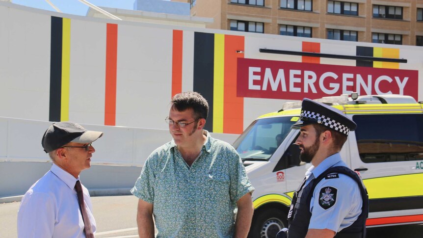 Two men and a police officer stand outside the emergency department of a hospital.