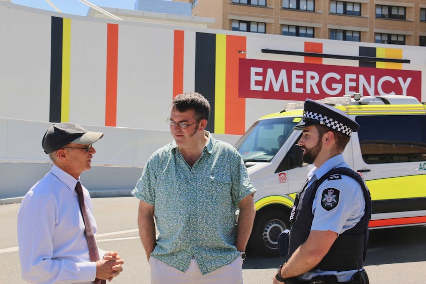 Two men and a police officer stand outside the emergency department of a hospital.