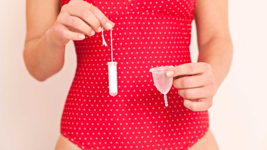 Midshot of a woman in red spotted underwear holding a menstrual cup in one hand and a tampon in the other.