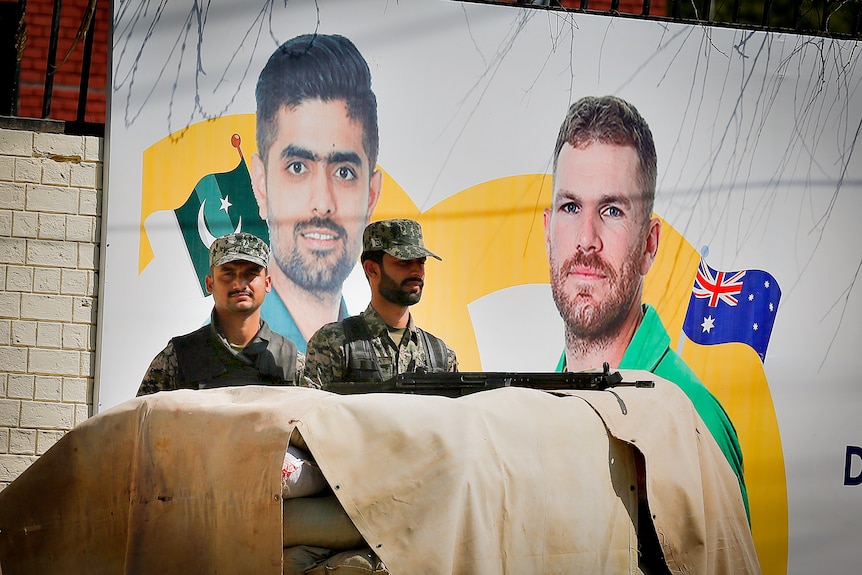 Two men in camo gear stand in front of a poster featuring the Pakistani and Australian cricket captains