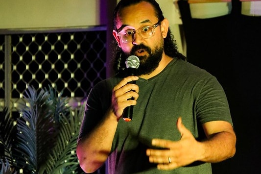 Bearded Indigenous male comedian performing stand-up. He is wearing an olive green T-shirt and is carrying a microphone