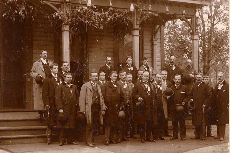 A black and white photo of men in suits standing outside a house
