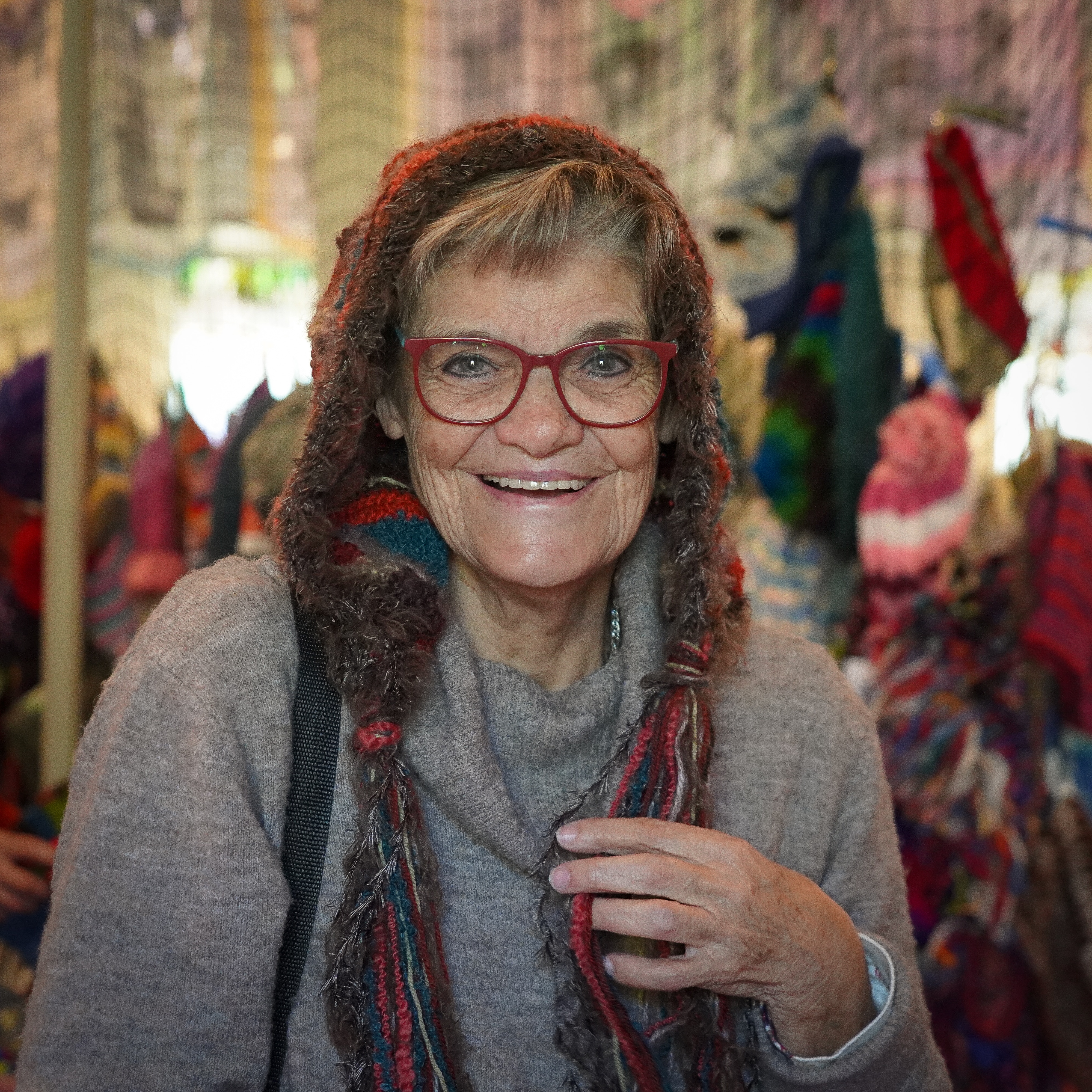 An older woman in reading glasses smiles at the camera, a knitted beanie rests on her head and cascades down over her shoulders.
