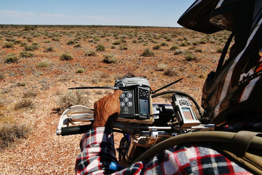 An Aboriginal man wearing a helmet sits on a motorbike looking at a GPS in the outback.