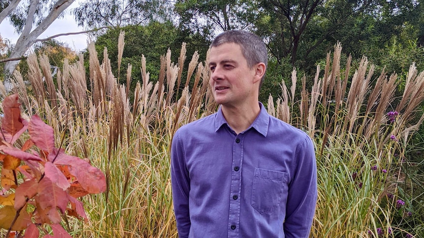 Simon Lush stands in grasslands with purple flowers and trees in the background, bright red-leafed plants in the foreground.