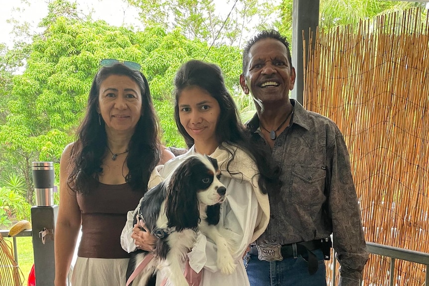 Destiny stands in between her parents while holding a dog and smiling. 