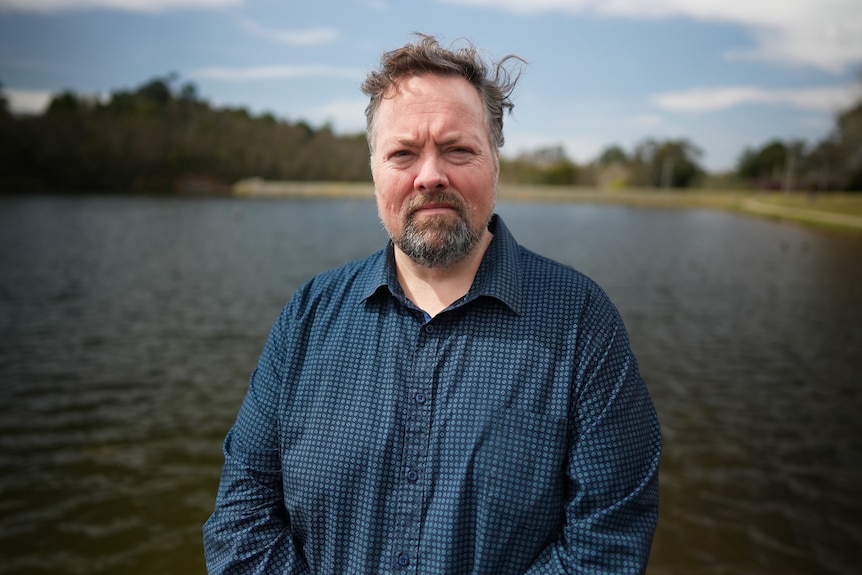 A middle-aged man with a greying beard stands in front of a small dam.