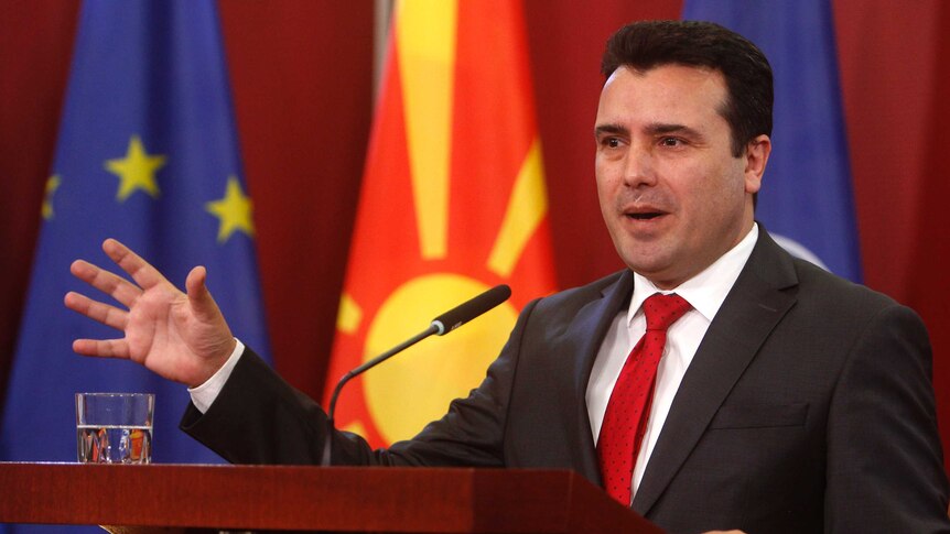 In front of NATO, EU and the Macedonian flag, Prime Minister Zoran Zaev gesticulates while talking behind a plinth.