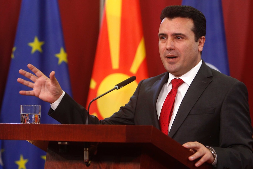 In front of NATO, EU and the Macedonian flag, Prime Minister Zoran Zaev gesticulates while talking behind a plinth.