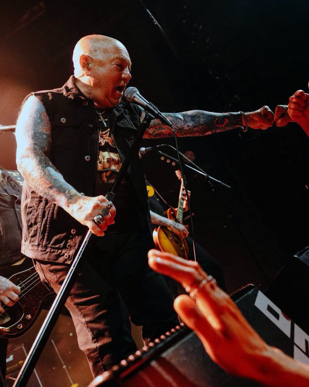 A bald man with tattooed arms in a black sleeveless shirt holds a microphone stand in hand and stretches the other arm out.