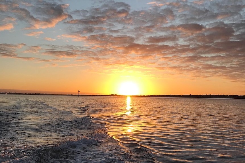 An orange sunset reflecting over a boat's wake spreading across a lake.