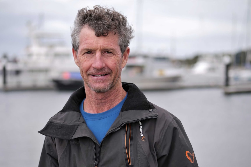 A middle-aged man stands in a marina infront of the ocean and ships.