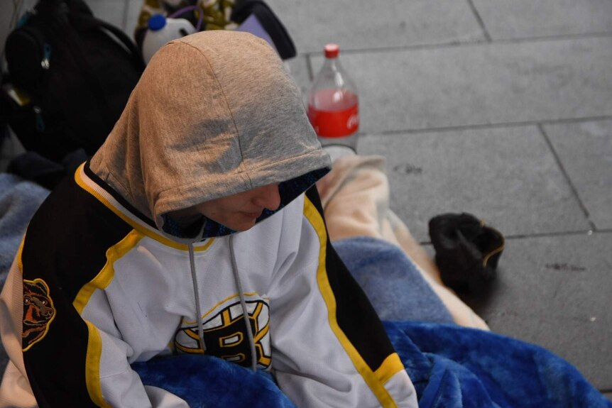 A homeless man sits on a blanket on a footpath wearing a hoodie.