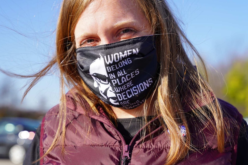 a woman with red hair and a black and white mask with text and a picture of Ruth Bader Ginsburg on it looks at the camera