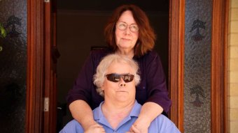 A woman rests her arms on the shoulders of a man in the wheelchair in front of her.