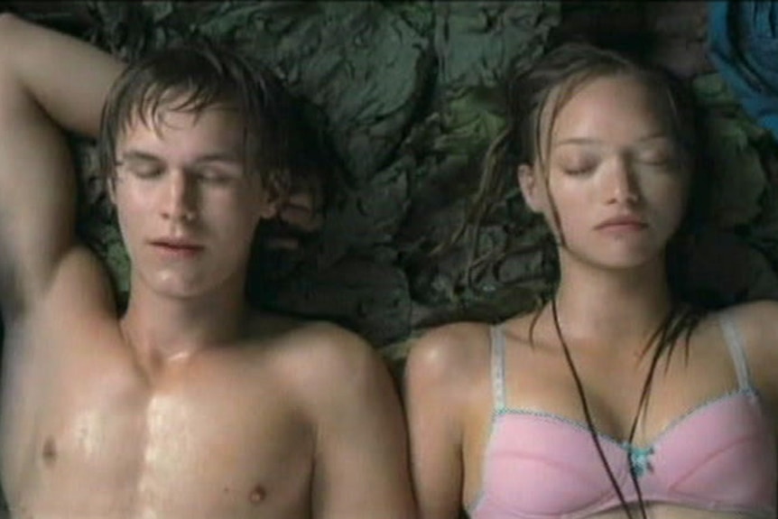 A young man with his shirt off and a woman in a pink bra lie next to each other.