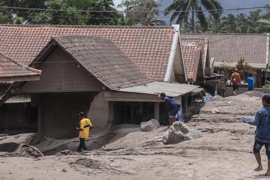 People walk along a street and pass houses covered by volcanic material.