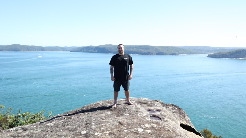 A man in black clothes stands on a rock with an expansive view of water and coast behind him