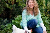 Jessamy Miller kneels down next to four chickens; two white, one spotted white and black and one black, in a leafy garden.