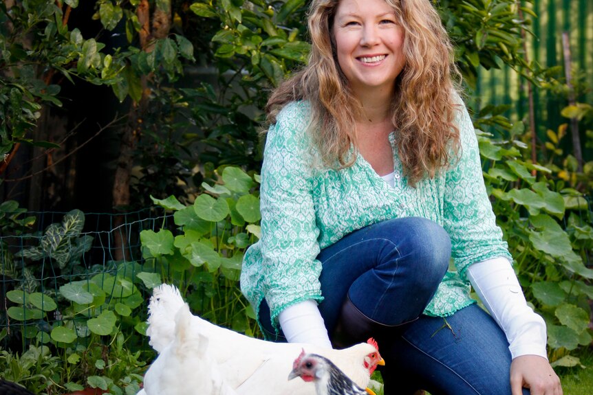 Jessamy Miller kneels down next to four chickens; two white, one spotted white and black and one black, in a leafy garden.
