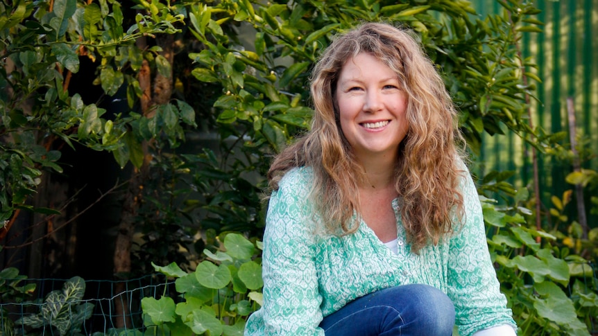 Jessamy Miller kneels down next to four chickens; two white, one spotted white and black and one black, in a leafy garden.