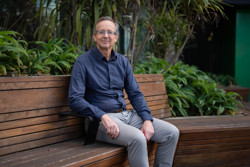 An older man wearing glasses sits on a bench in a garden and smiles