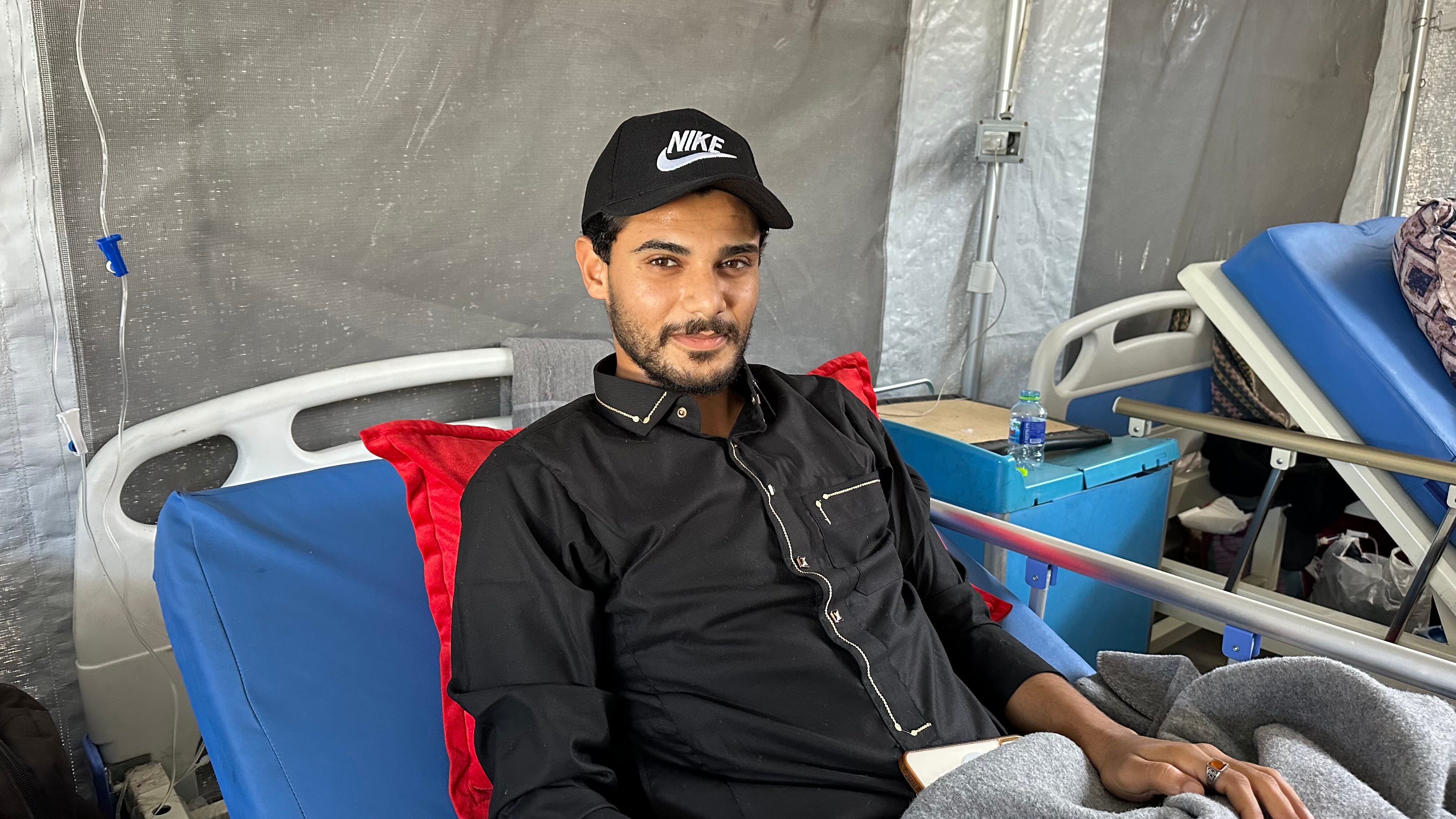 A Palestinian man in black shirt and Nike cap sits on a hospital bed 