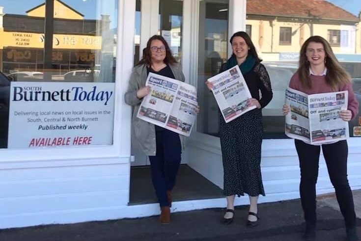 Three women holding newspapers standing outside a white building.