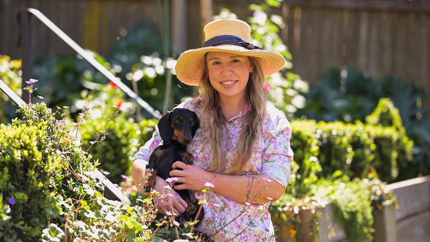 Woman with long hair and floral dress standing in garden smiling, holding pet dachshund dog.